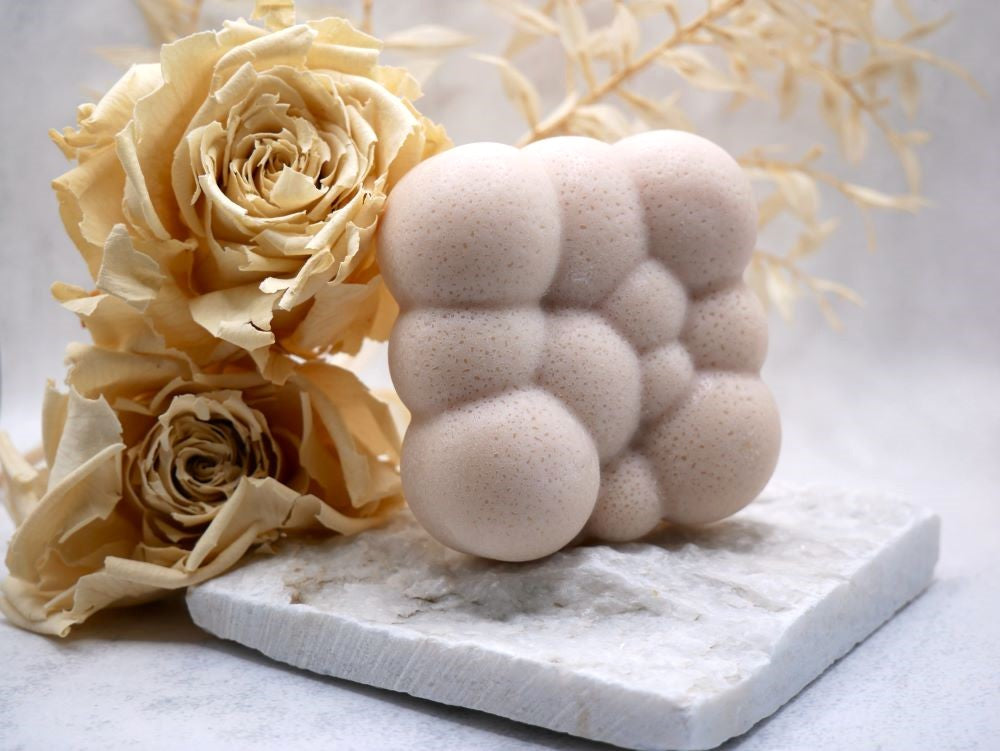 Pina Colada Seife in Steinoptik von Wertvoll Naturkosmetik & Seifenmanufaktur. Die Seife steht auf einer weißen unebenen Steinplatte, dahinter zwei beige Trockenblumen (beige Rosen). Der Hintergrund ist ebenfalls beige- grau gehalten.