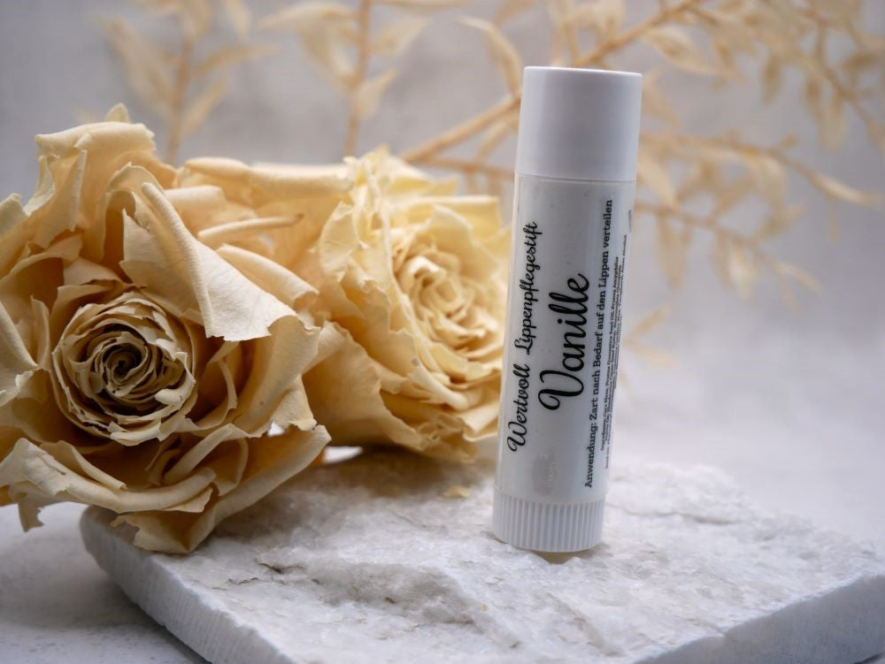 Lippenpflegestift von Wertvoll Naturkosmetik & Seifenmanufaktur mit der Aufschrift Vanille. Er steht auf einer weißen unebenen Steinplatte, dahinter zwei beige Trockenblumen. Der Hintergrund ist ebenfalls beige- grau gehalten.