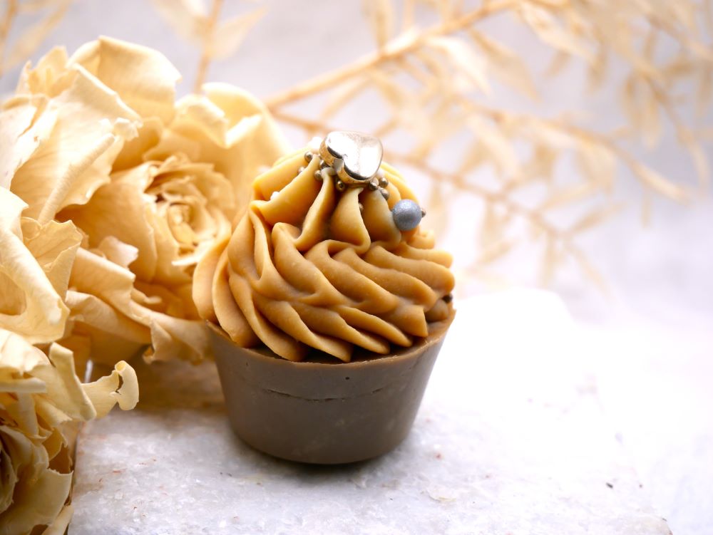 Ein kleiner Seifencupcake mit grauem Töpfchen und cremefarbener Sahnehaube mit Zuckerstreuseln  von Wertvoll Naturkosmetik & Seifenmanufaktur. Im Hintergrund sind verschiedene Trockenblumen in beige.