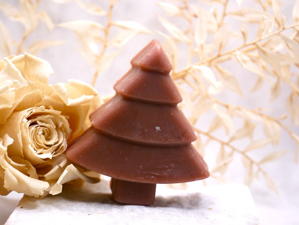 Apfel- Zimt Seife von Wertvoll Naturkosmetik & Seifenmanufaktur.  Tannenbaum Seife mit Ornamenten und mit brauner Farbe, sie steht auf einer weißen Steinplatte. Im Hintergrund sieht man beige Trockenblumen.
